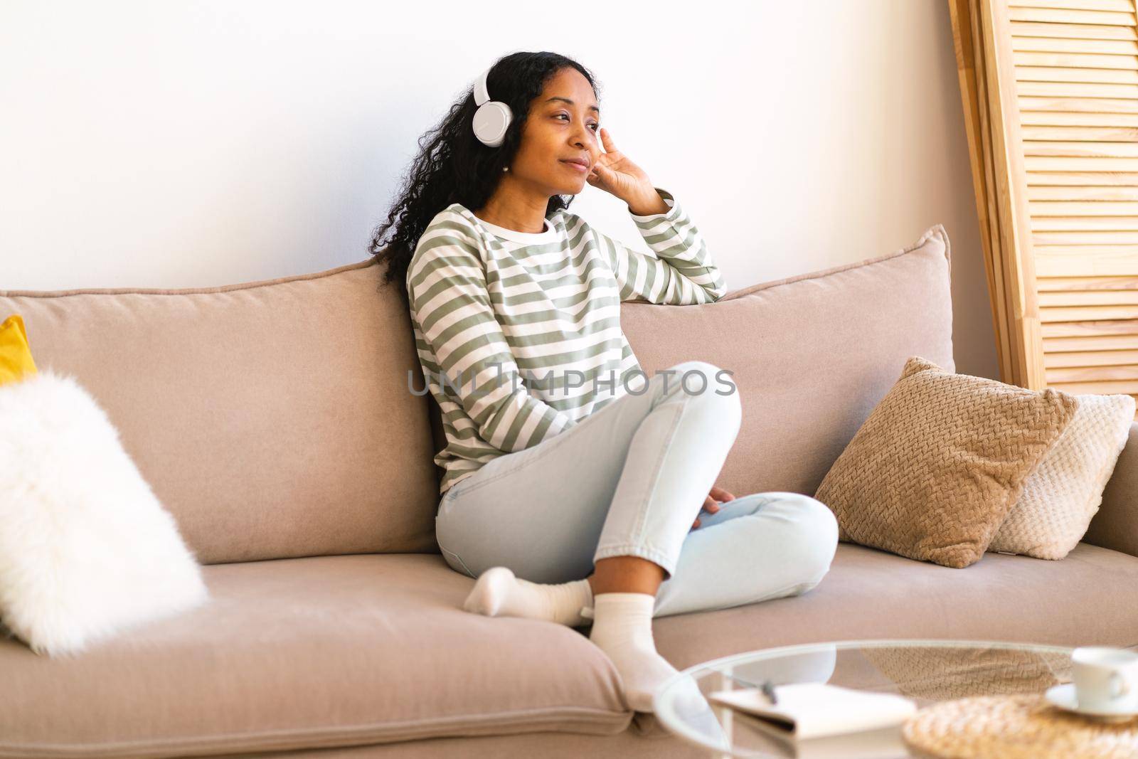 Smiling african-american female listening to audiobook in headphones while sitting on coych in living room. Concept of mind escaping and self-help trend. Staying at home. Horizontal, copy space