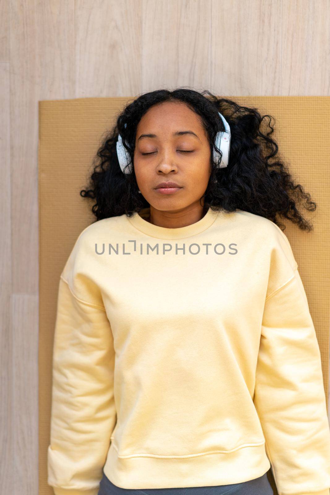 African-american female in sportswear lying on mat with closed eyes. Wearing headphones and listening to music while having rest after training. Vertical of concentrated woman relaxing