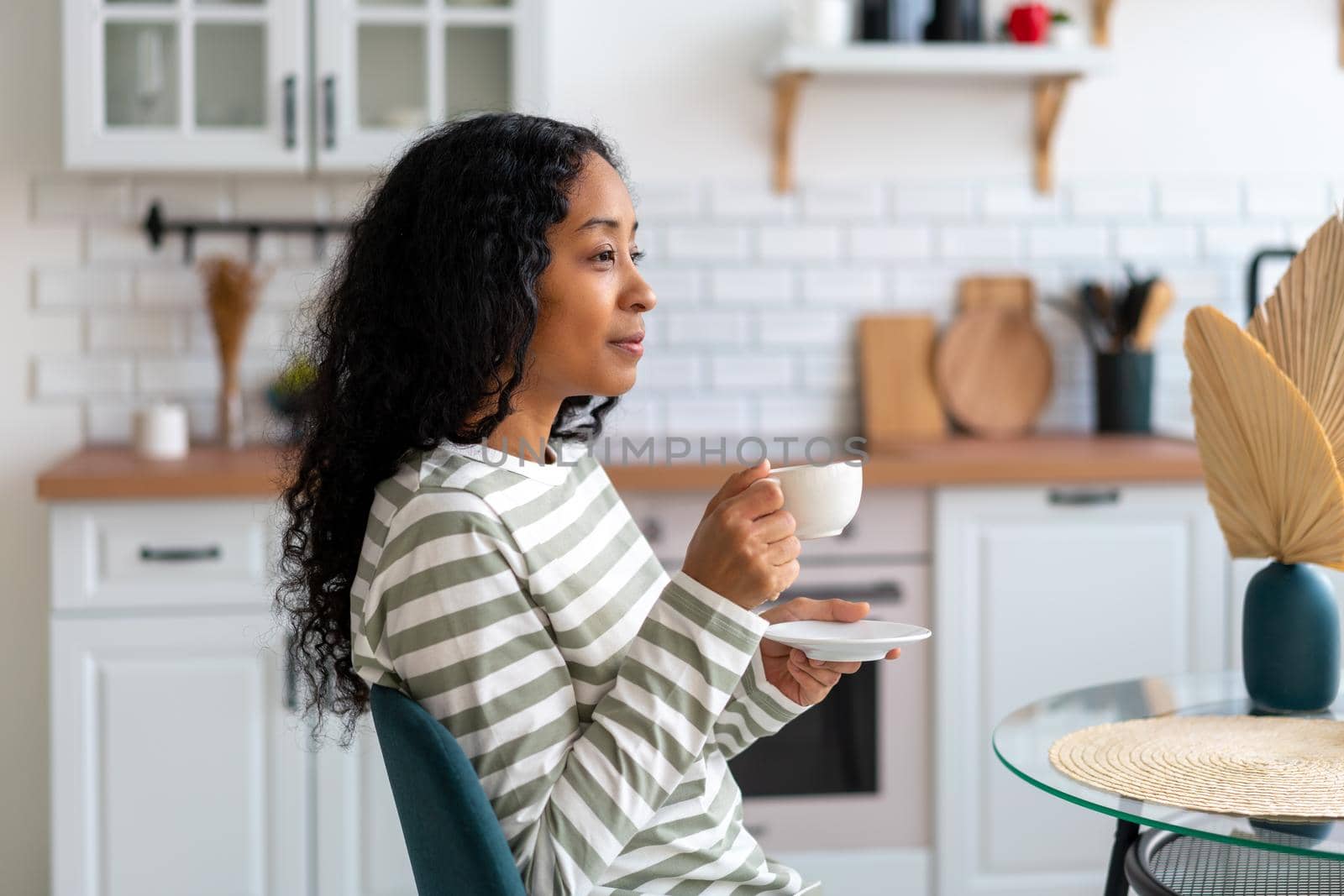 African-american woman drinking cup of coffee in the morning for breakfast. Concept of niksen lifestyle and slow-living. Modern white kitchen on background. Small break, micro-moment