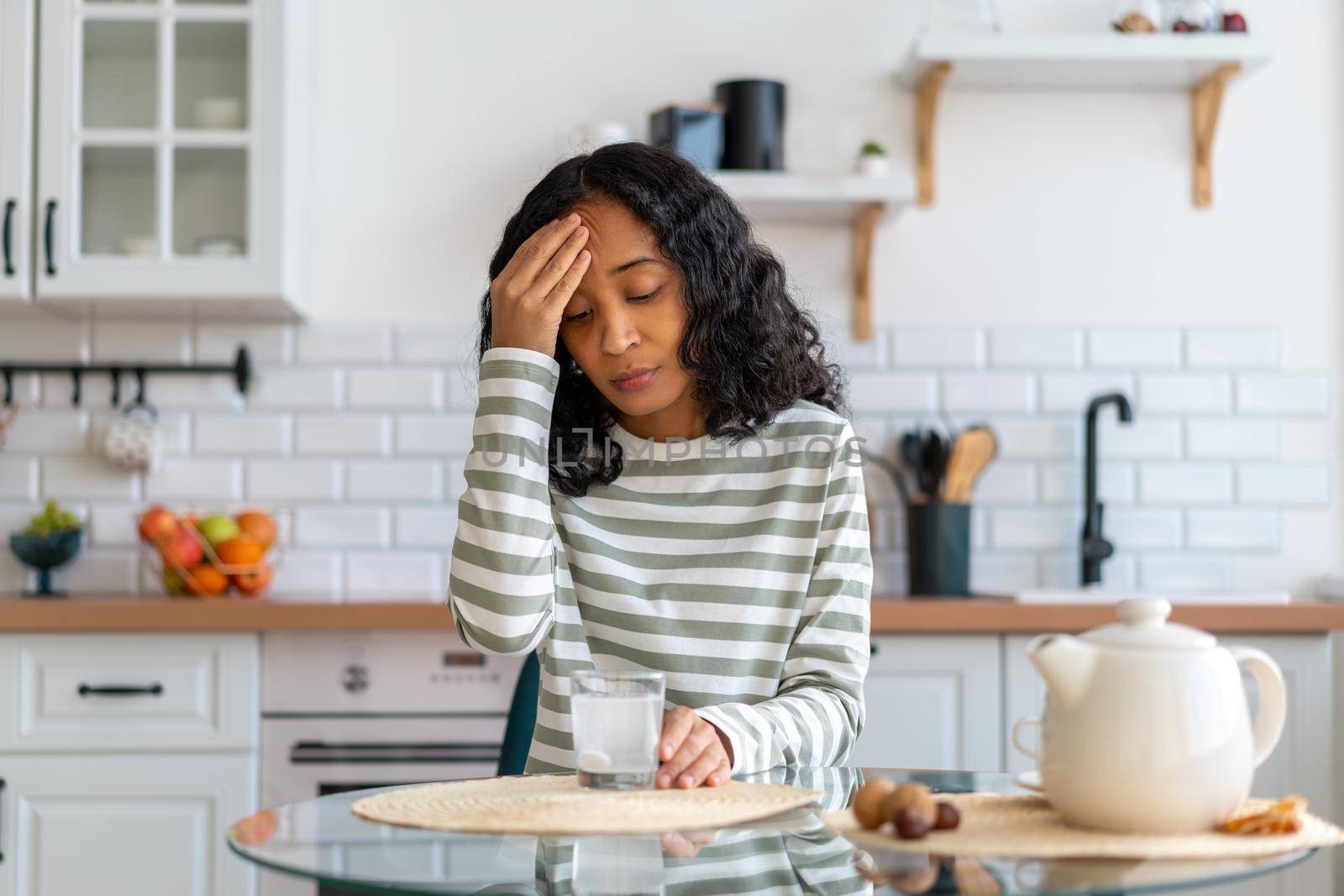 Concept of african-american woman having headache. Dissolving fizz drug in glass of water in kitchen. Taking painkiller to cure pain. Feeling tired and exhausted due to sickness. Touching forehead