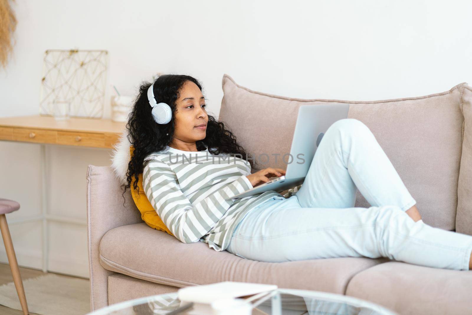 African-american female in headset chatting online on laptop while lying on sofa in living room by NataBene