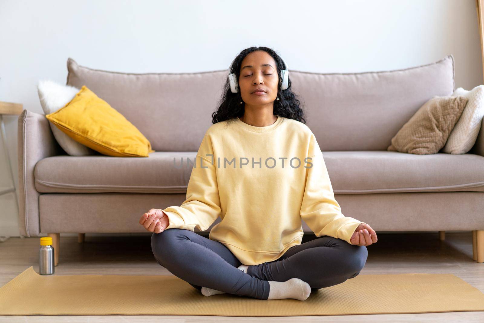 Young african-american female in headphone listening to music while meditating on mat in living room by NataBene