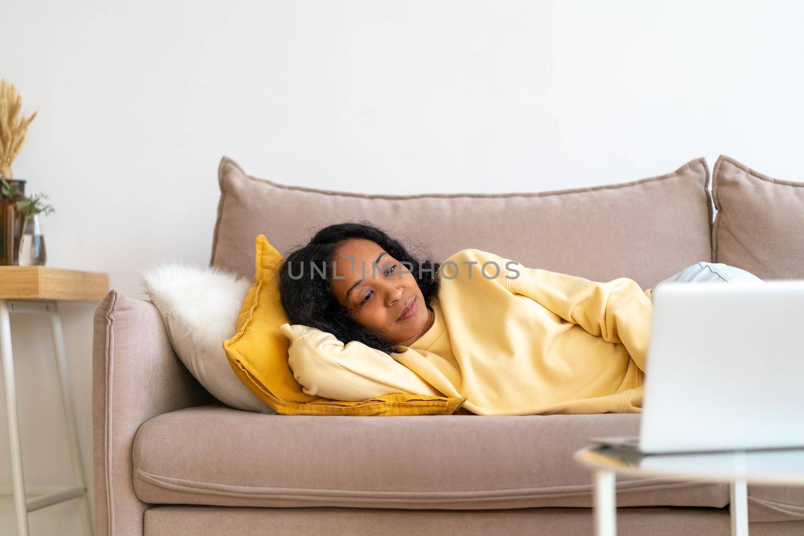 African-american woman watching films on laptop while lying on couch in living room. Concept of spending free time at home. Entertainment for millennials. Relaxing on quarantine due to covid