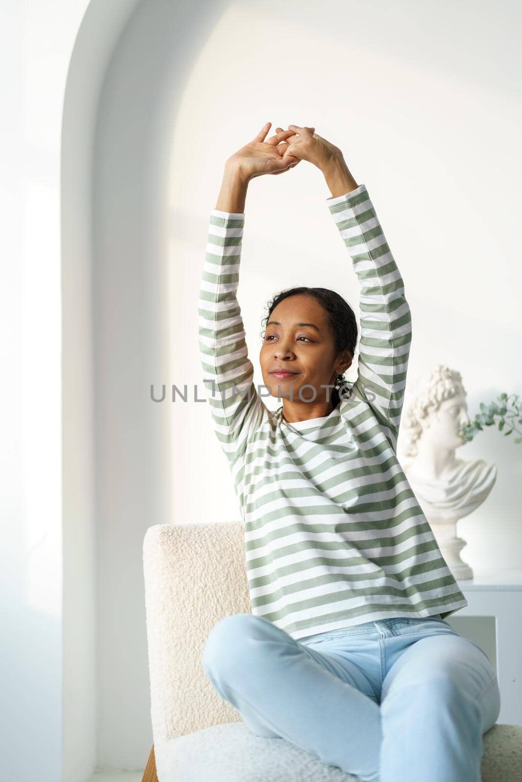 African-american woman in casual stretching while sitting on chair. Calm looking at window in morning. Concept of gathering thoughts together. Enjoying time alone. Happy smiling
