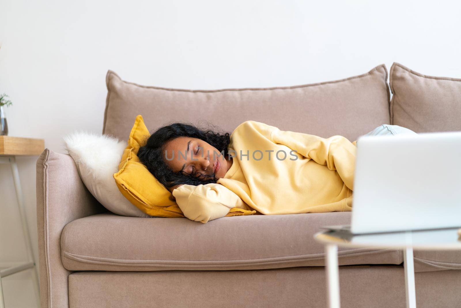 Sleepy young african-american lying and sleeping on sofa in living room while movie playing on laptop. Boring spending free time watching film. Concept of snackable wellness and micro-moment