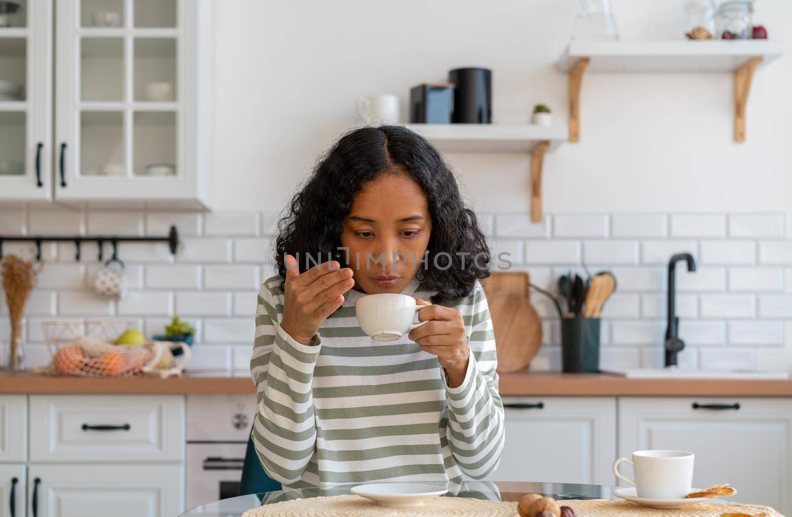 African-american female having long-term covid effect. Concept of loosing sense of smell. Lack of coffee flavor. Coronavirus symptoms. Sitting in white kitchen. Dealing with long-lasting blindness