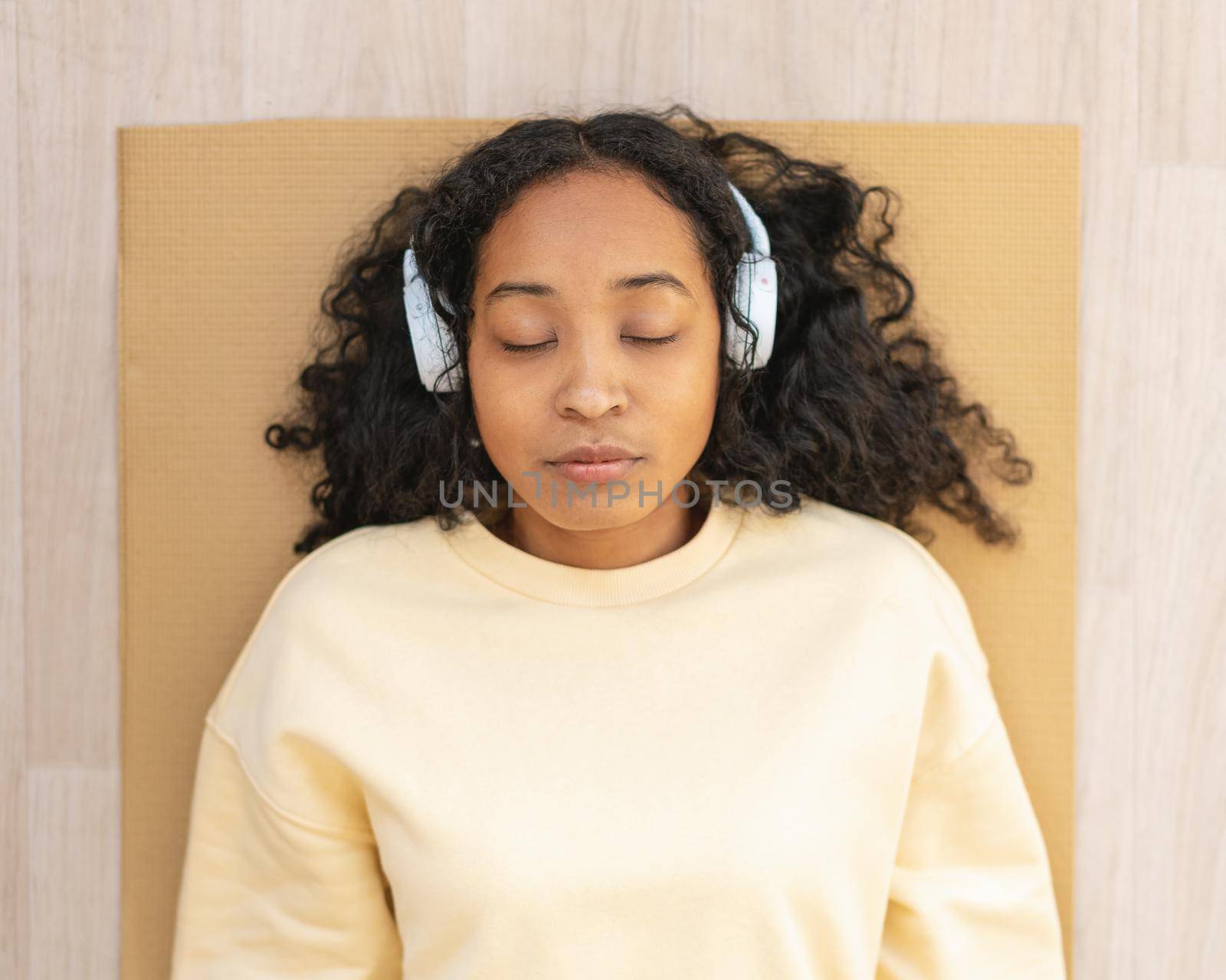Tired and sleepy african-american female lying on mat. Wearing earphones and listening to music while having rest after training. Horizontal of woman relaxing, closeup. Meditation practice