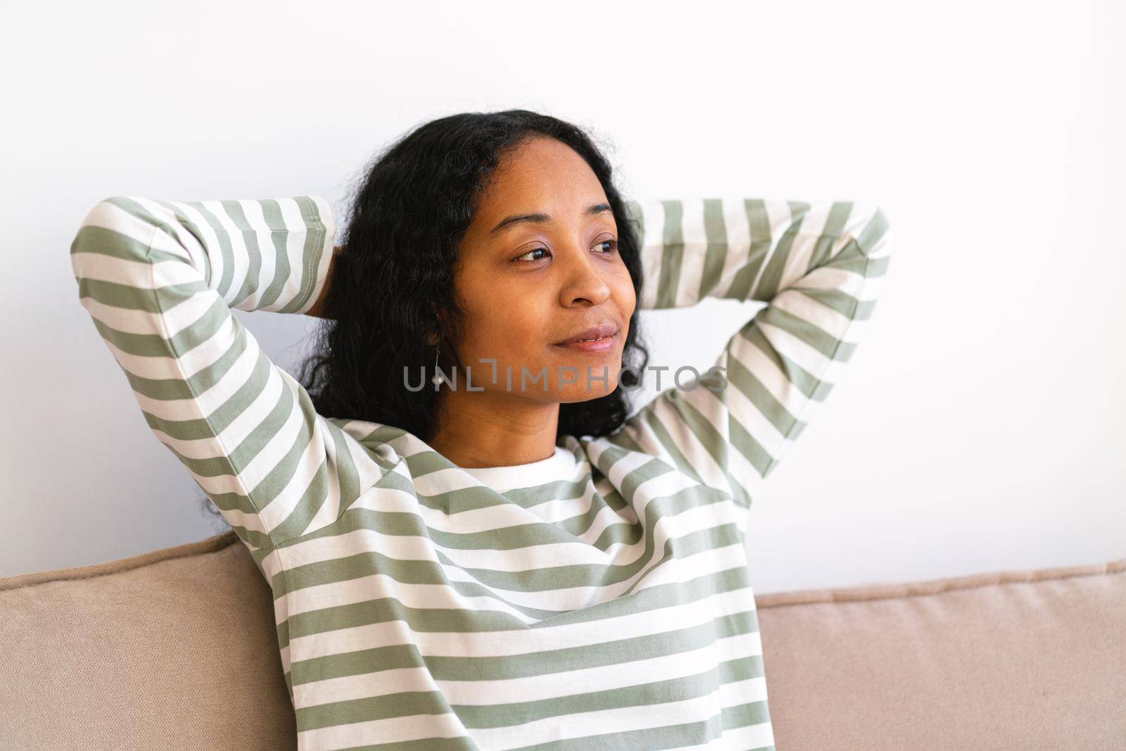 Happy young african-american female putting hands behind head while sitting on couch in living room. Concept of enjoying life and slow pace lifestyle. Thoughtful smiling woman self-reflecting