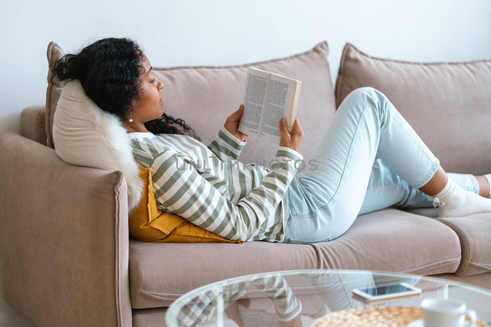 Faceless intelligent frican-american female lying on couch and reading book. Taking technology break and limiting screen time. Healthy digital break. Having micro-moment and enjoying life