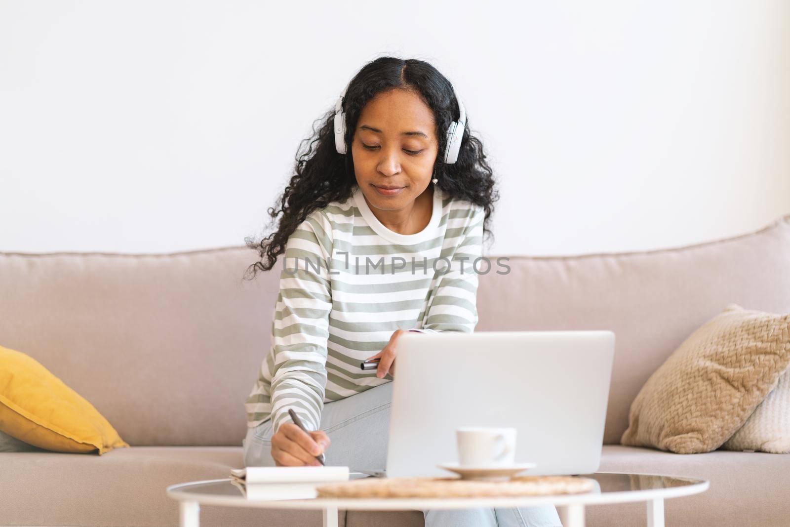 Young-african american student making notes while listening to online lecture on laptop. Concept of wireless e-learning at home. Focused woman in headset studying on sofa. Concept of multi-tasking