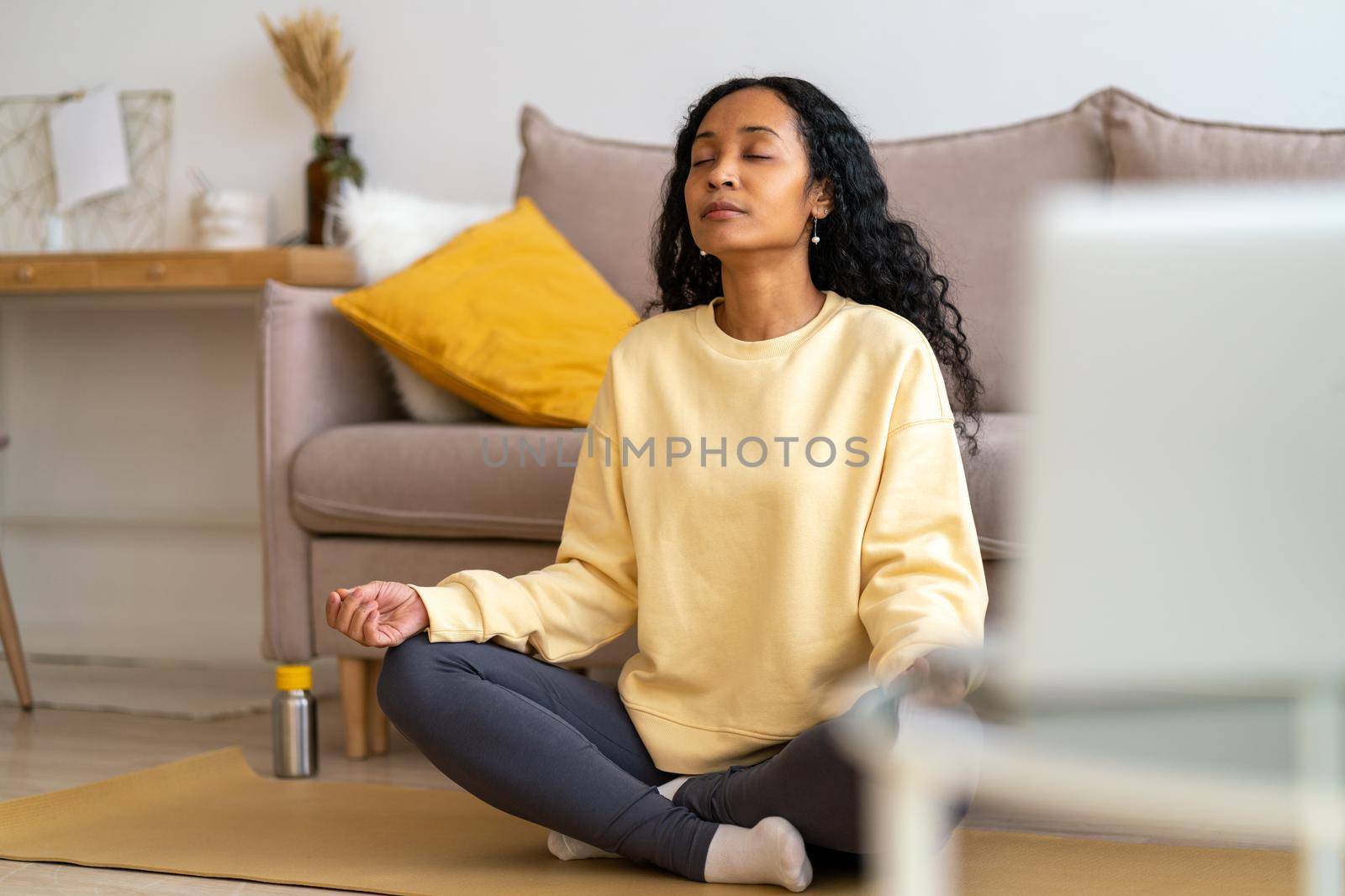 Young african-american female sitting in lotus pose on mat in living room while meditating by NataBene