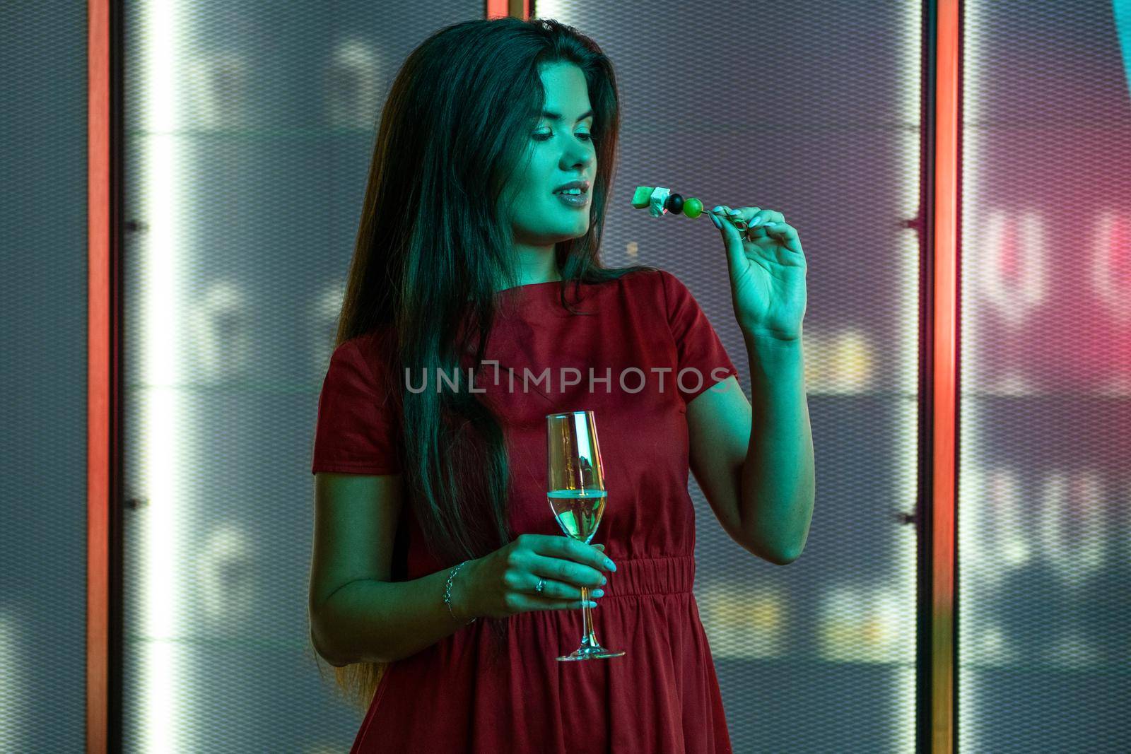 Young long-haired brunette in red dress enjoying champagne with light snacks in nightclub while standing alone in subdued color lighting