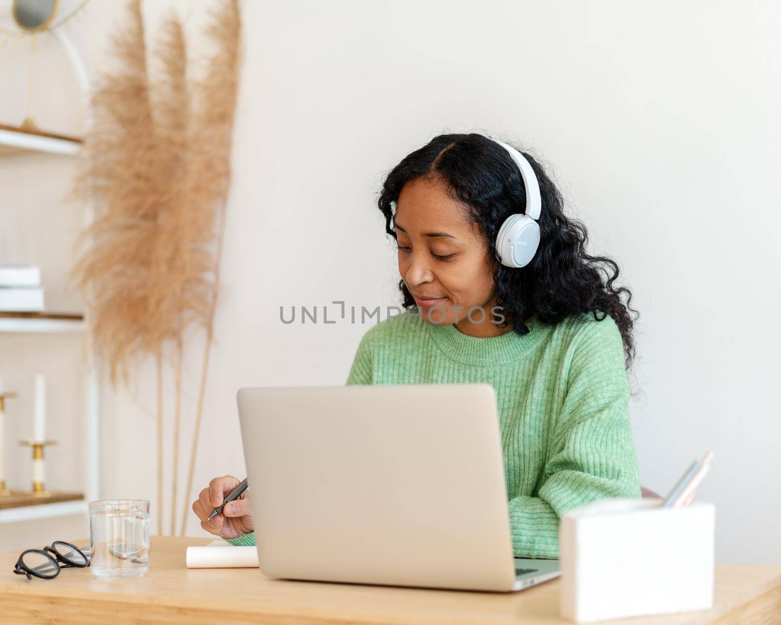 African-american female making notes in notebook while listening to audio podcast in earphones. Taking online course on laptop. Modern e-learning education. Young student distance studying