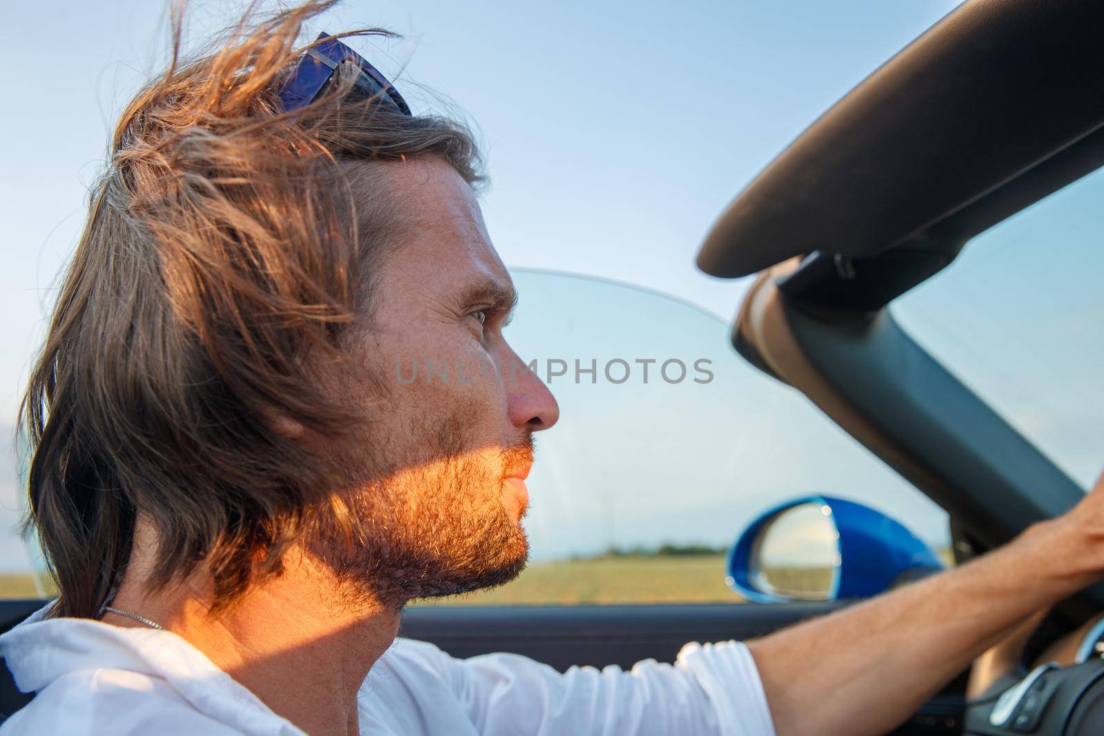 The handsome brutal man with long hair and an easy beard at a car without top at sunset, cabriolet, blue color, sunglasses by vladimirdrozdin