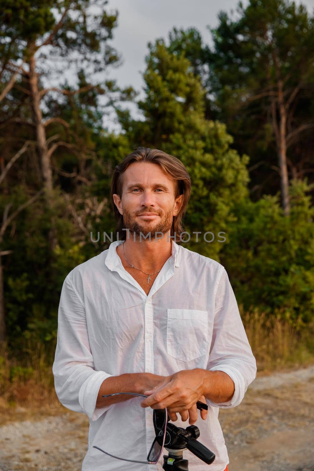 Portrait of handsome brutal man with long hair at sunset, the blue eyes, an easy bristle, he is dressed in a white shirt with a short sleeve, green trees on a background. High quality photo