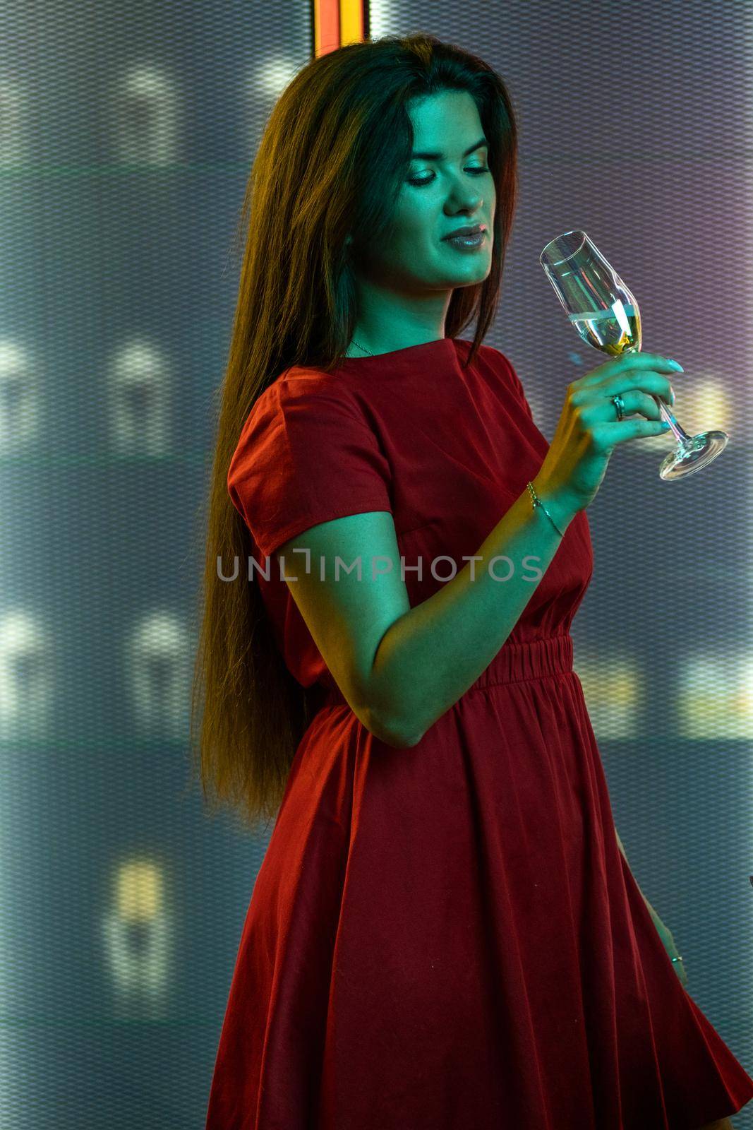 Young attractive brunette in red dress drinking champagne at party, standing alone in subdued color lighting holding glass