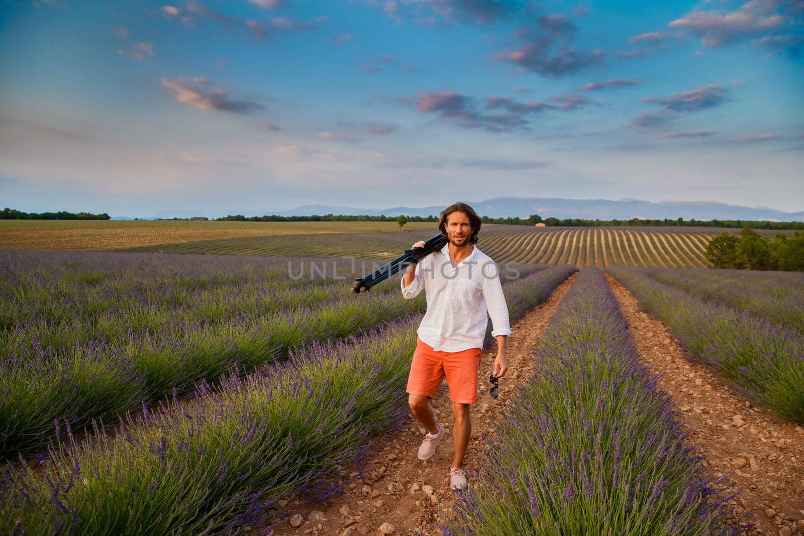 The handsome brutal man with long brunette hair is walking with tripod in the field of lavender in provence near Valensole, France, clear sunny weather, in a rows of lavender, red shorts, white shirts. High quality photo