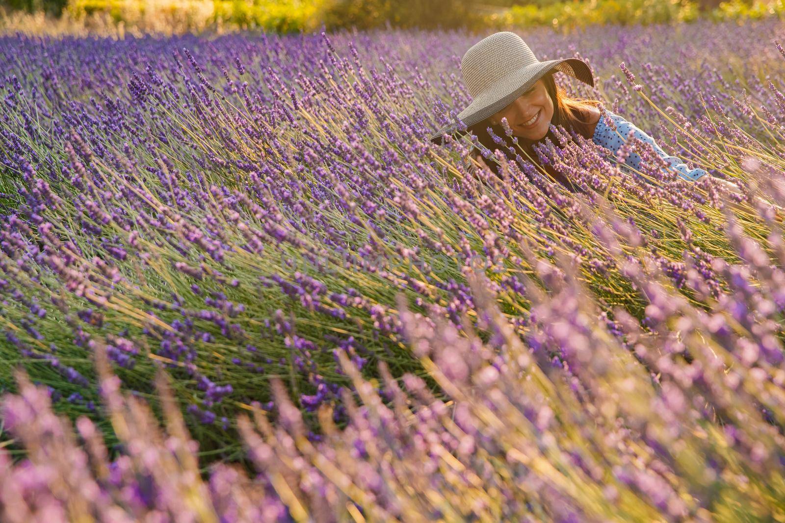 The beautiful young girl in a blue dress and cap sits in a lavender flowers, long curly hair, smile, pleasure, a house of the gardener in the background, trees, perspective of a lavender. High quality photo