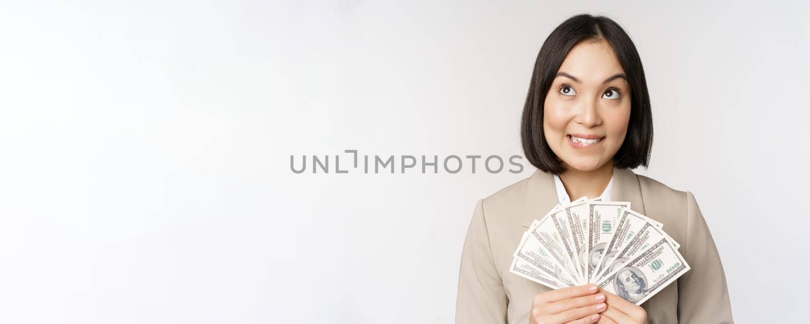 Image of asian corporate woman, happy businesswoman showing money, cash dollars and thinking, standing in suit over white background by Benzoix