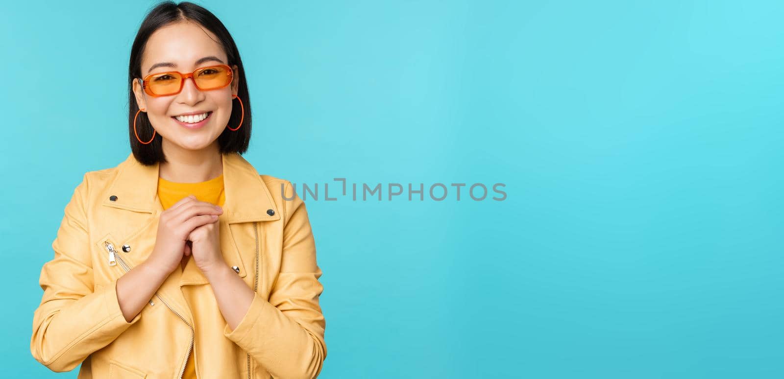 Portrait of asian woman in sunglasses, looking hopeful, flattered, smiling happy, standing over blue background. Young people concept