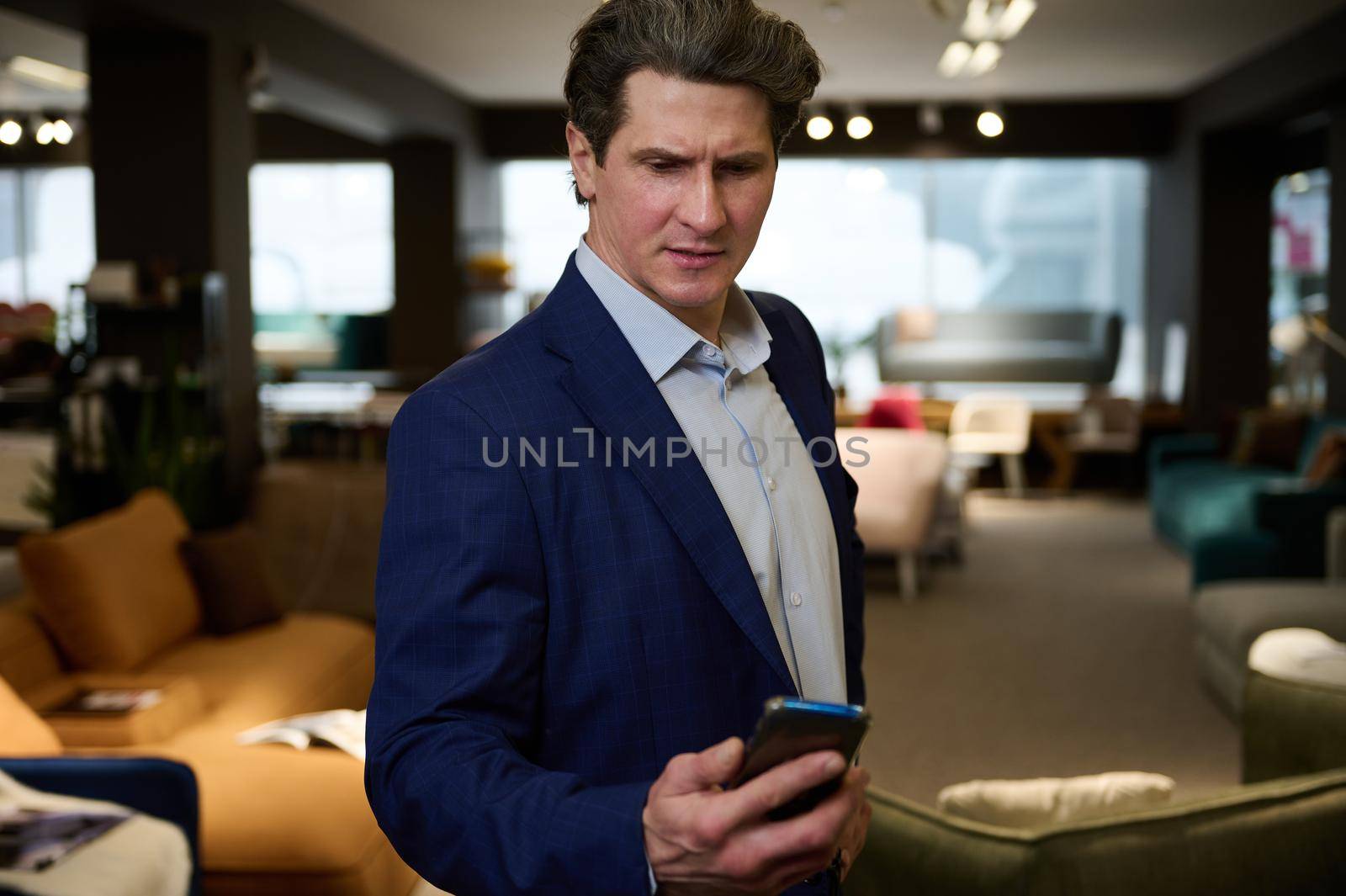 Busy retail assistant, successful sales manager, confident business owner, entrepreneur, handsome imposant Caucasian man in business suit holds mobile phone in the furniture store showroom