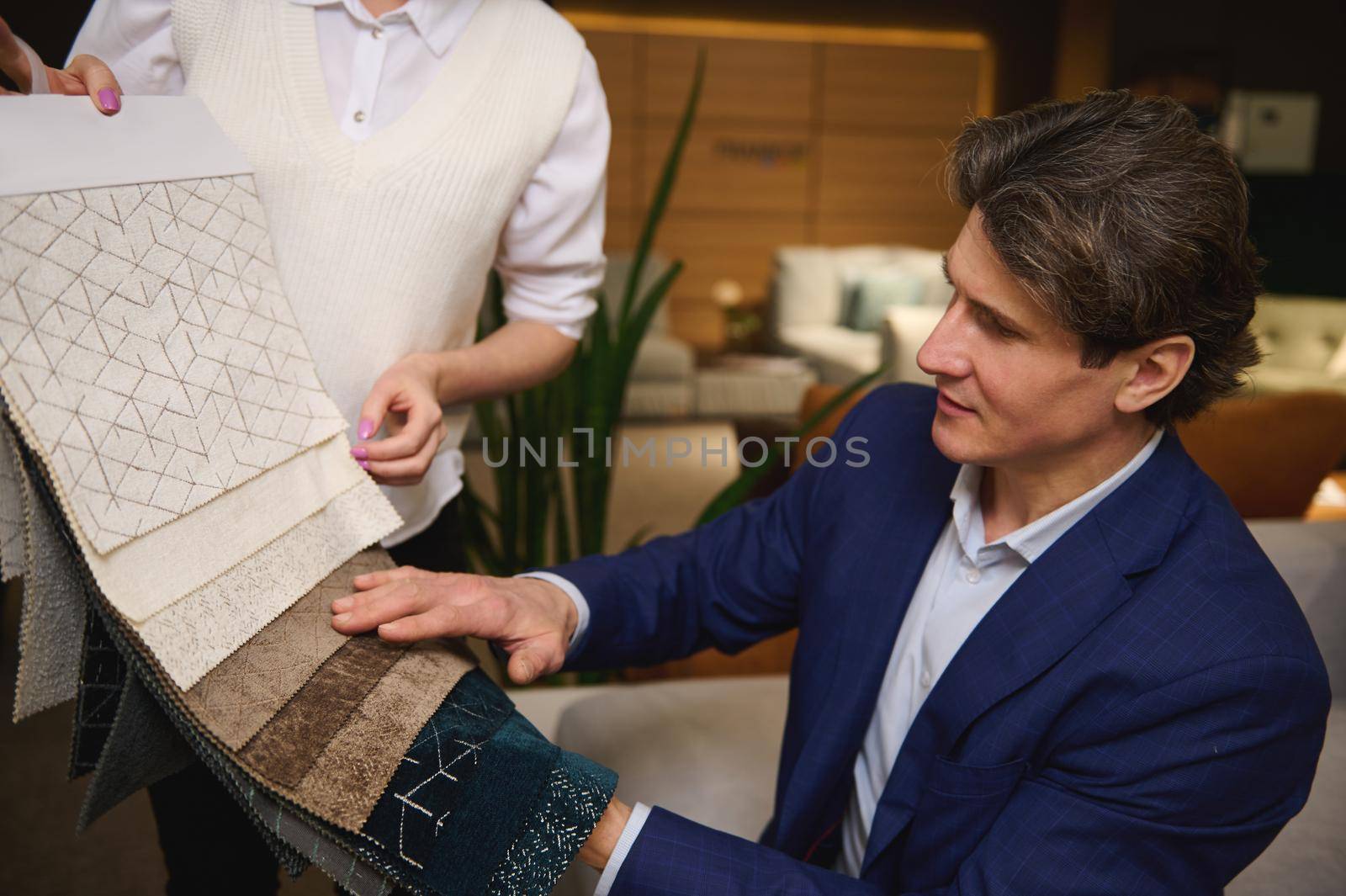 Charming middle aged man, interior designer in business suit puzzled by the choice of upholstery material for a new sofa, examines fabric samples in the hands of a sales assistant in a furniture store