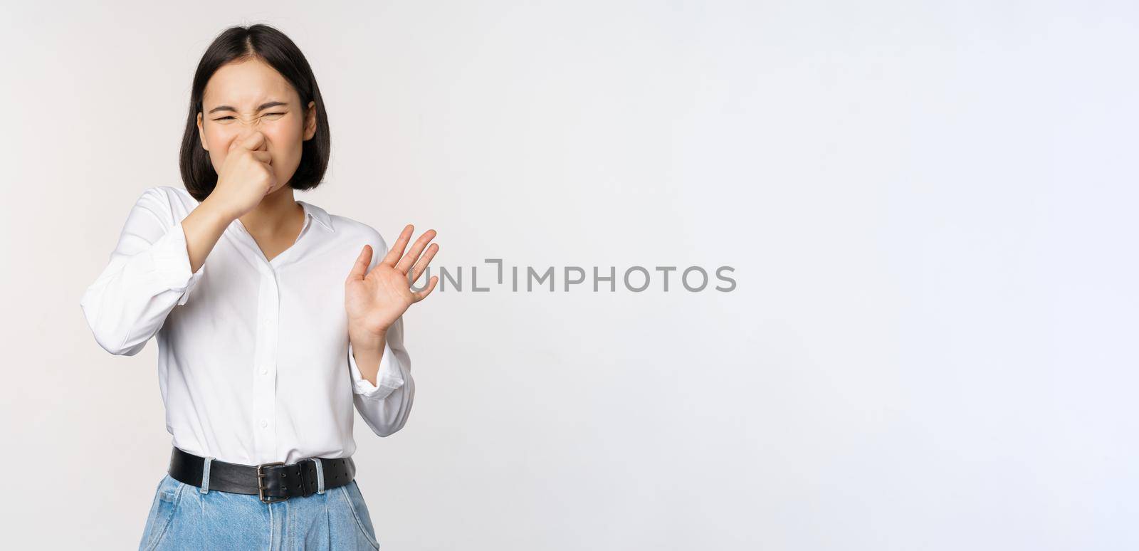 Disgusted asian girl close nose and showing rejection gesture, digusting bad smell, standing over white background by Benzoix