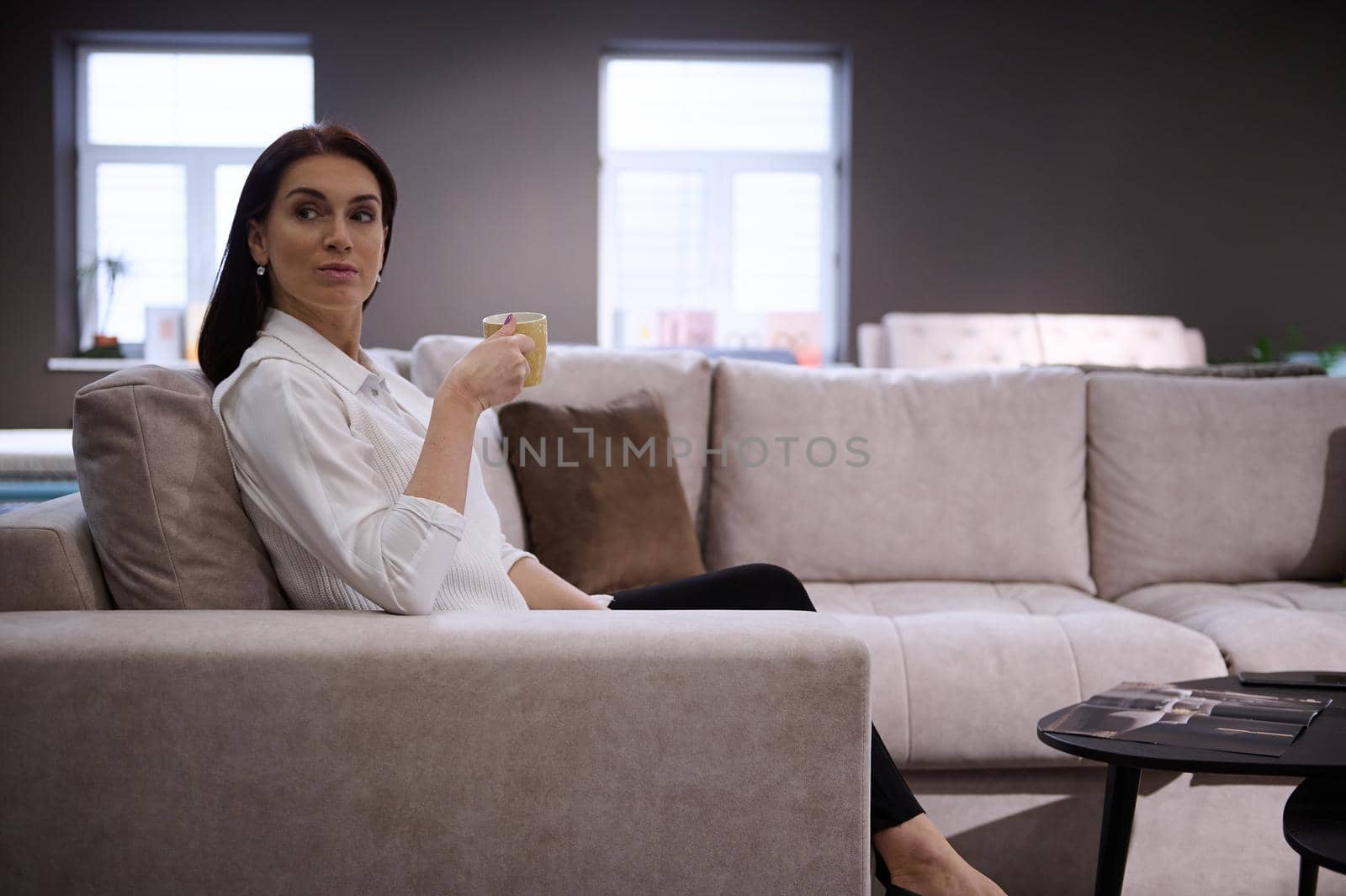 Relaxed beautiful European woman drinking coffee sitting on the comfortable sofa in the furniture store showroom by artgf