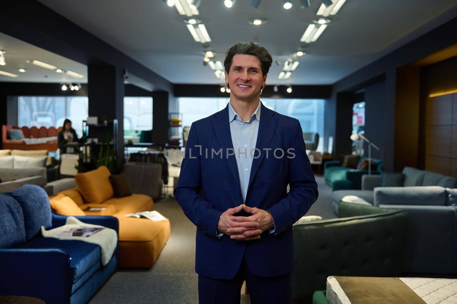 Pleasant kind handsome mature Caucasian man, confident business owner, retail assistant , sales manager in the showroom of a furniture store with wide range of sofas and upholstered furniture