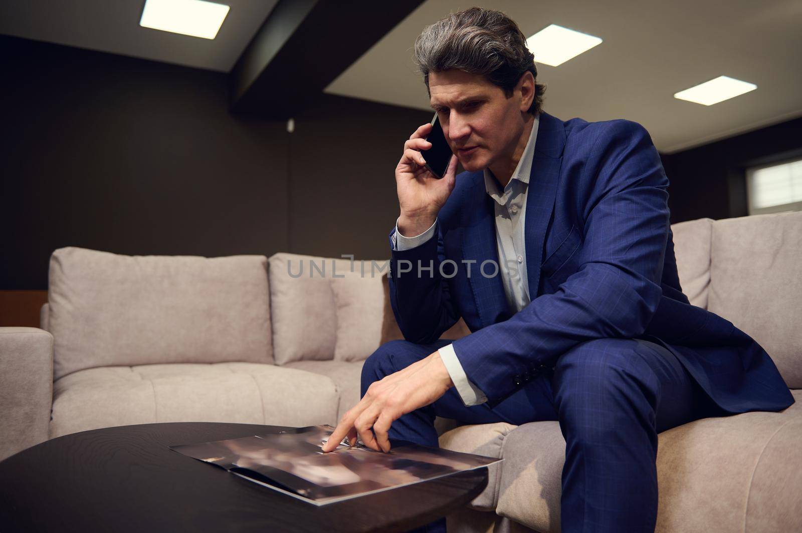 Handsome imposing mature Caucasian man, interior designer, successful businessman, entrepreneur, sales manager negotiating and advising clients on phone, looking furniture catalog in his cozy office