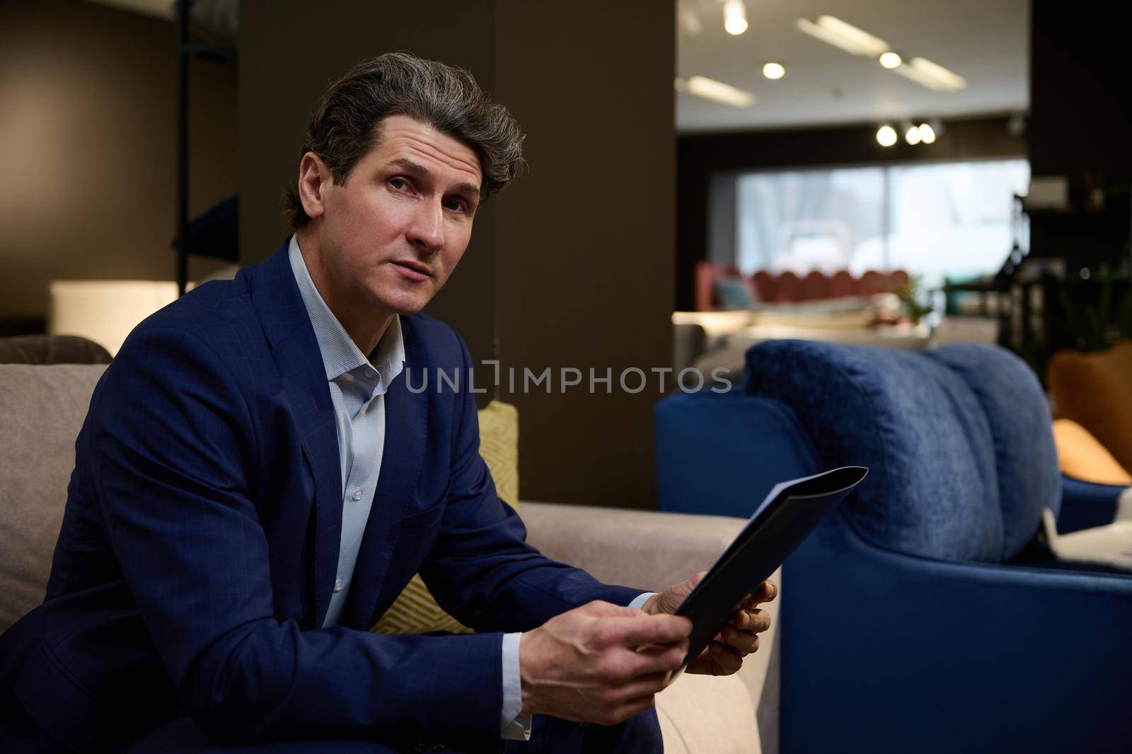 Charming mature Caucasian man with catalog sitting on a sofa in the furniture store and confidently looking at camera