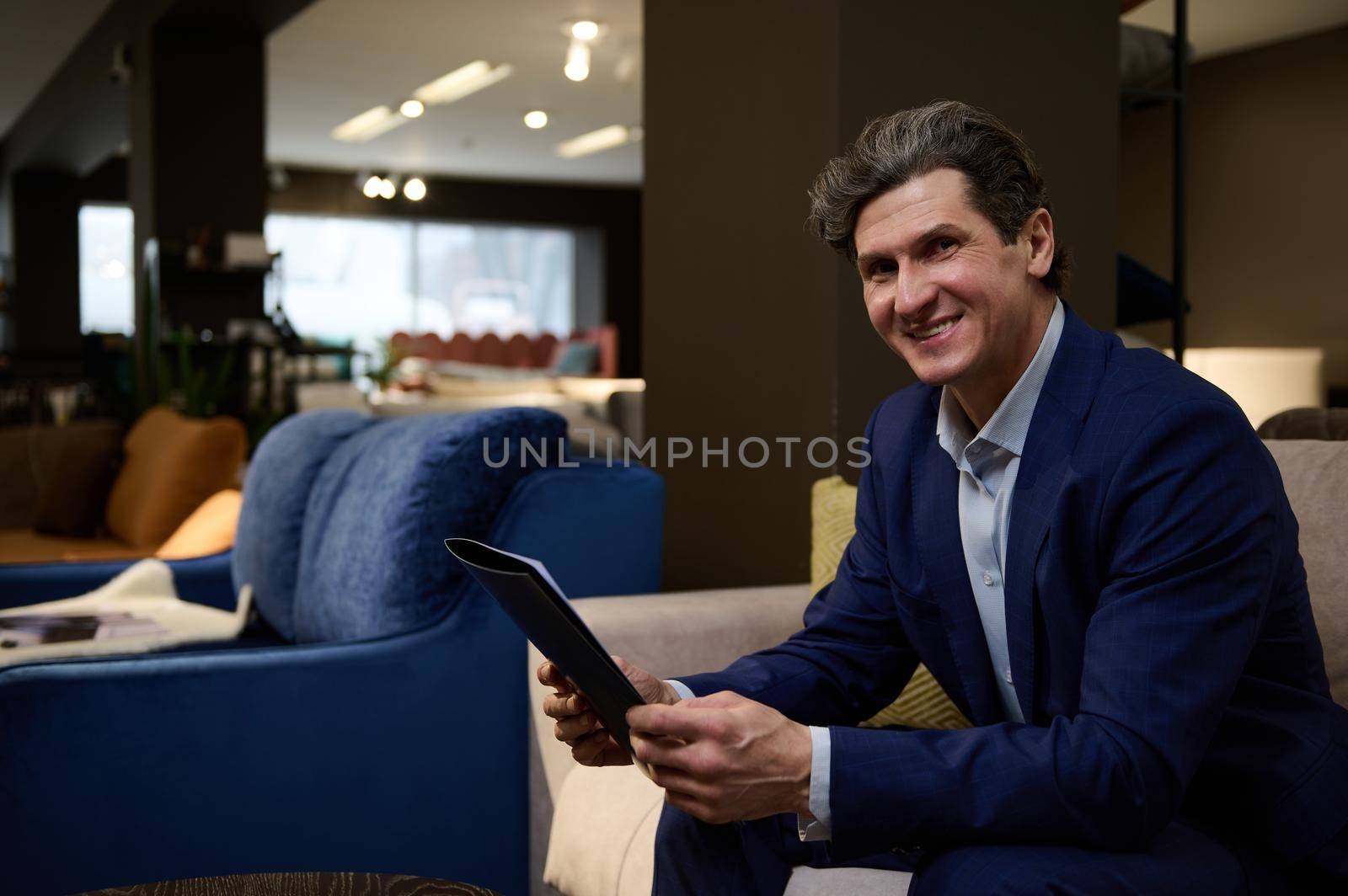 Charming mature Caucasian man with catalog sitting on a sofa in the furniture store and smiles toothy smile confidently looking at camera