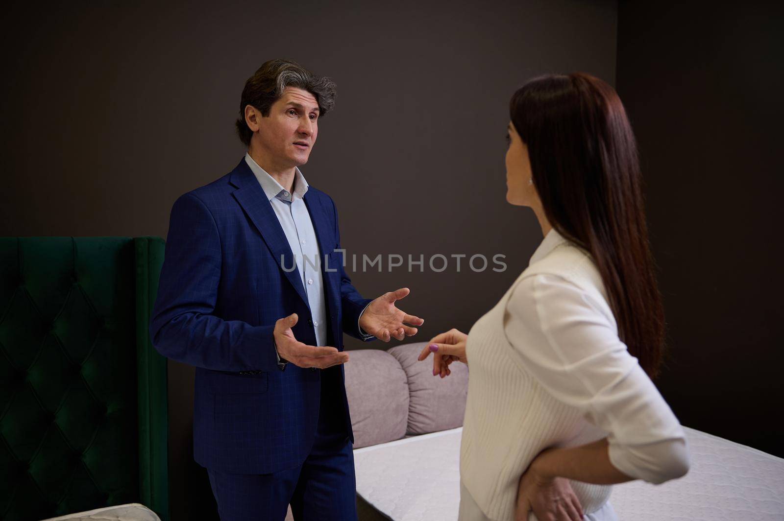 Successful sales rep, manager, assistant in upholstered furniture store showroom presents orthopedic mattresses to female customer, helping her to make purchase for home improvement. Interior design