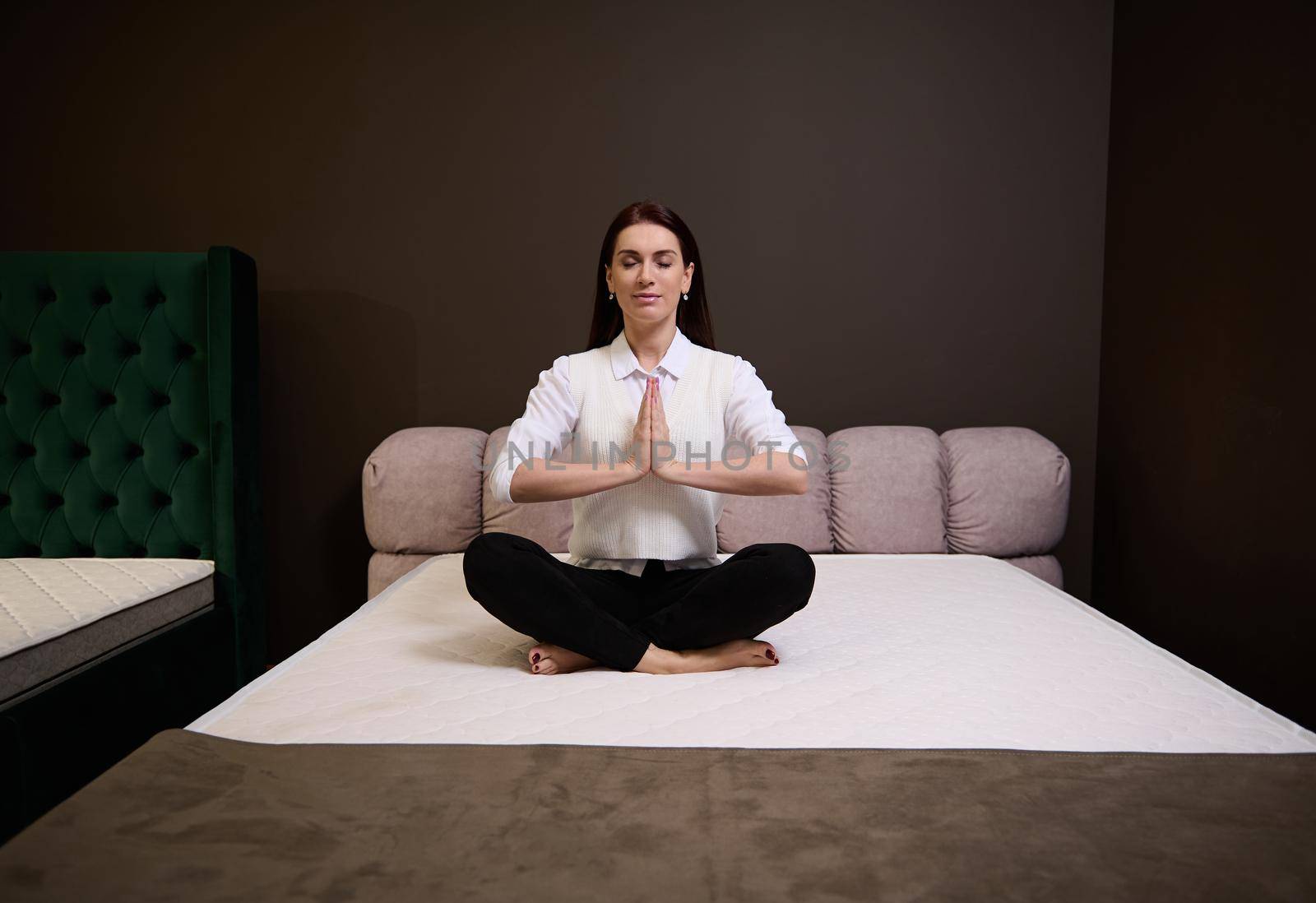 Serene woman meditating sitting in lotus position on an orthopedic mattress in the exhibition hall of a furniture store. Bedding accessories, beds for bedchamber by artgf