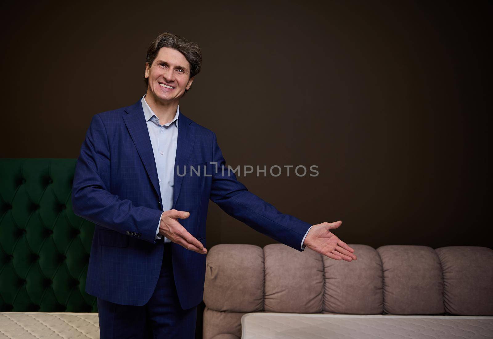 Handsome man sales manager consultant pointing at an upholstered bed and orthopedic mattress in the showroom of furniture store by artgf