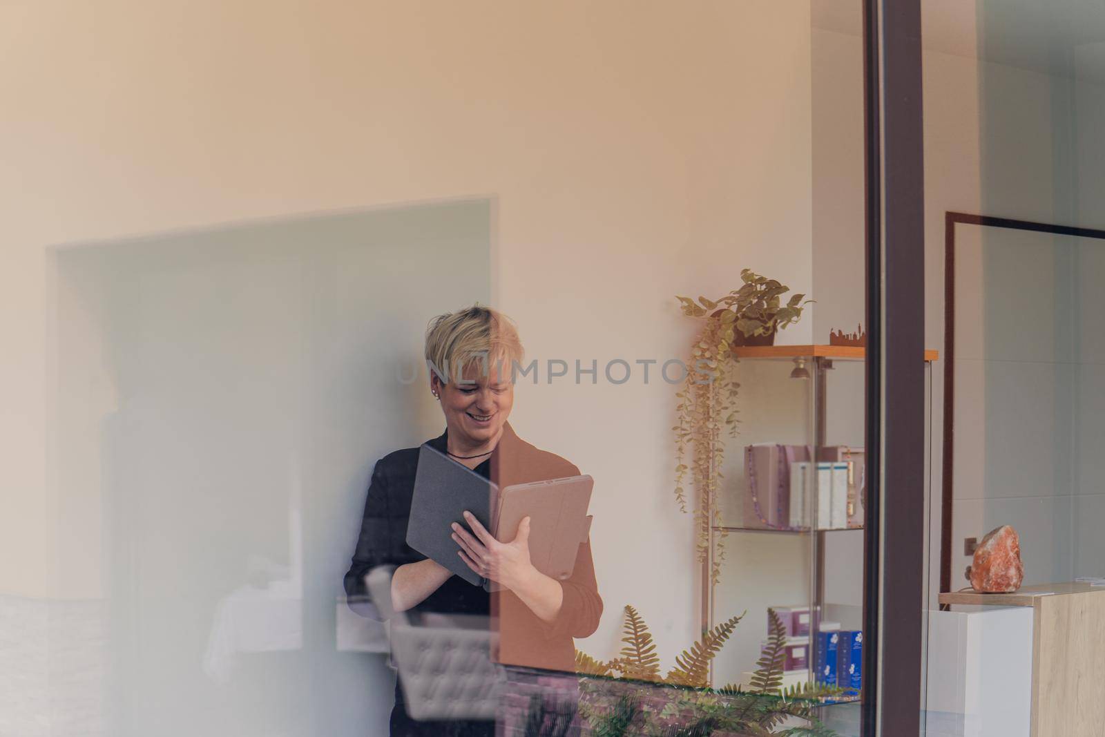 portrait through a window of a smiling mature professional Beautician with blonde hair, dressed in black work uniform, checking email on her Tablet. owner in her small beauty salon looking at laptop. Work from home, enterprising woman, active small business owner. Relaxed atmosphere and soft lighting from window, natural light, tablet. horizontal.