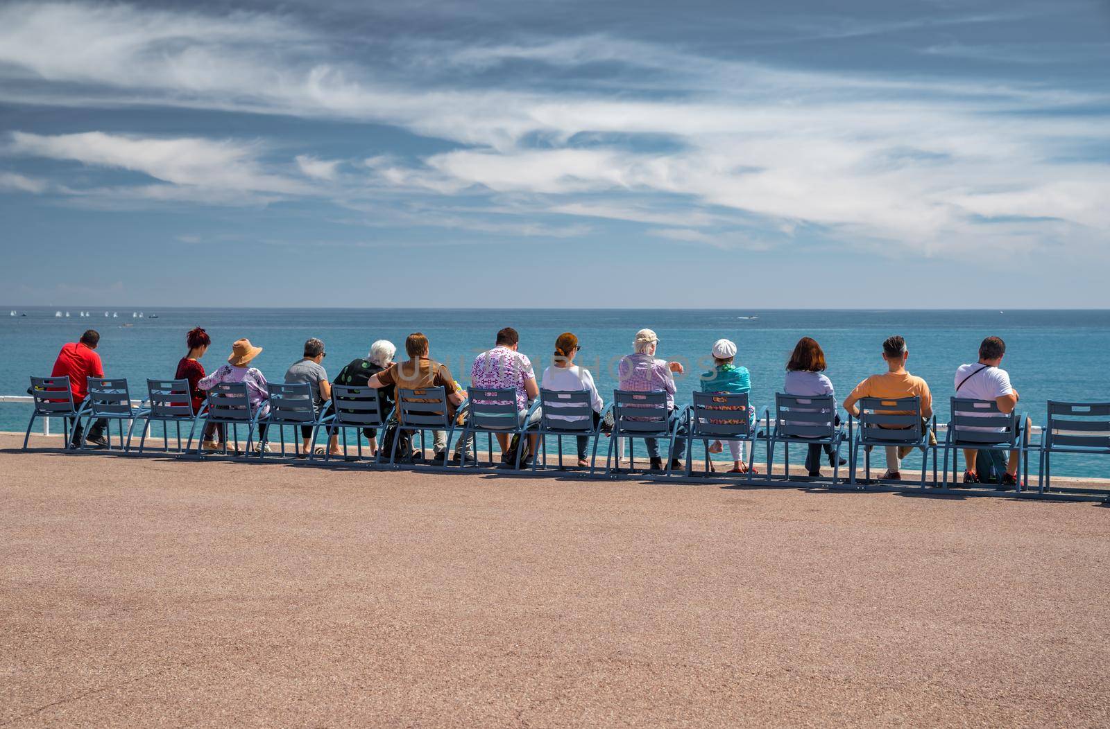 People sit on the famous blue chairs on embankment in Nice - France and watch at azure water of the Ligurian Sea, tranquillity, an old age, rest, age elderly people, the horizon, social. High quality photo