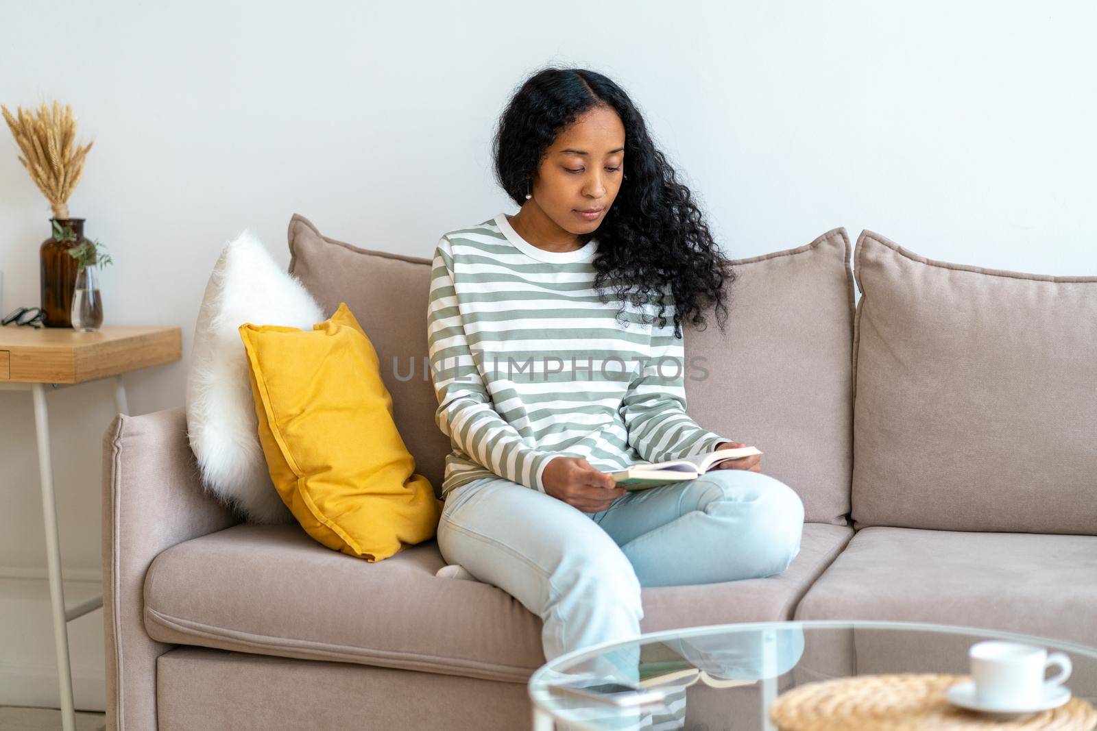 African-american woman in casual comfy clothing reading book while sitting on couch in cozy living room. Neutral tones interior. Concept of maintaining emotional wellbeing and tech independency