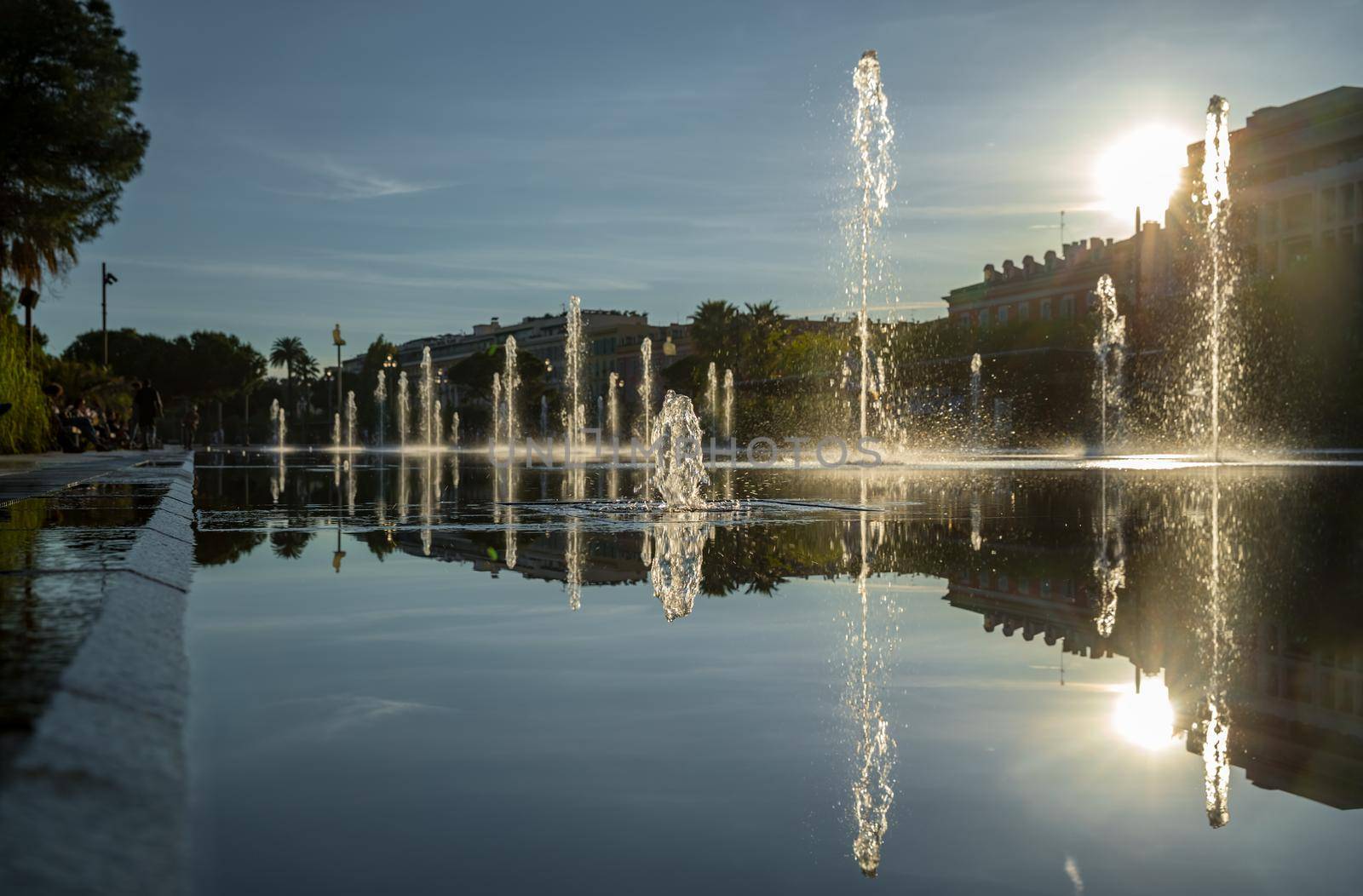 France, Nice, Famous place of Nice, square of fountain, mirror of water, playing kids, walking couples, a couple of old people are basking in the sunset, sun reflection, passing tram. High quality photo