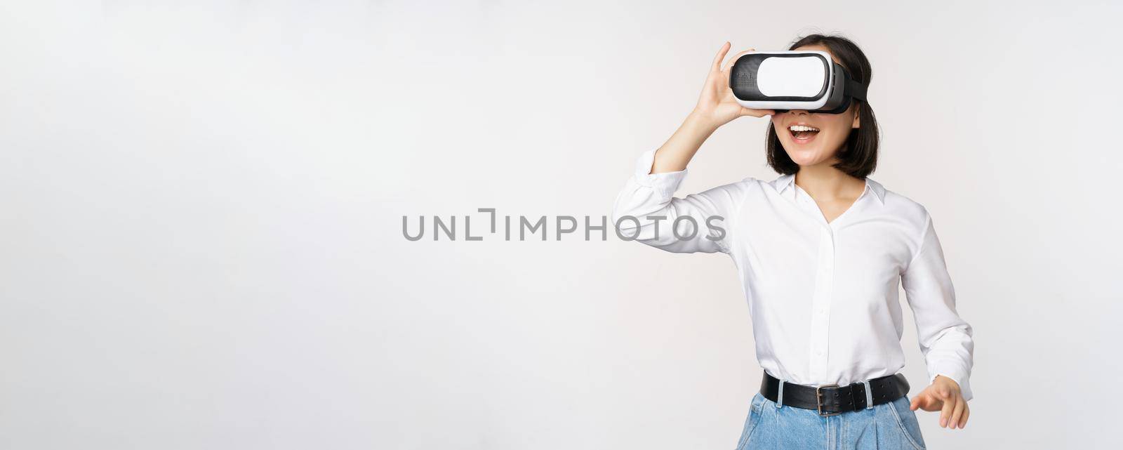 Excited young woman enter virtual reality in her glasses. Asian girl using vr headset, standing over white background by Benzoix