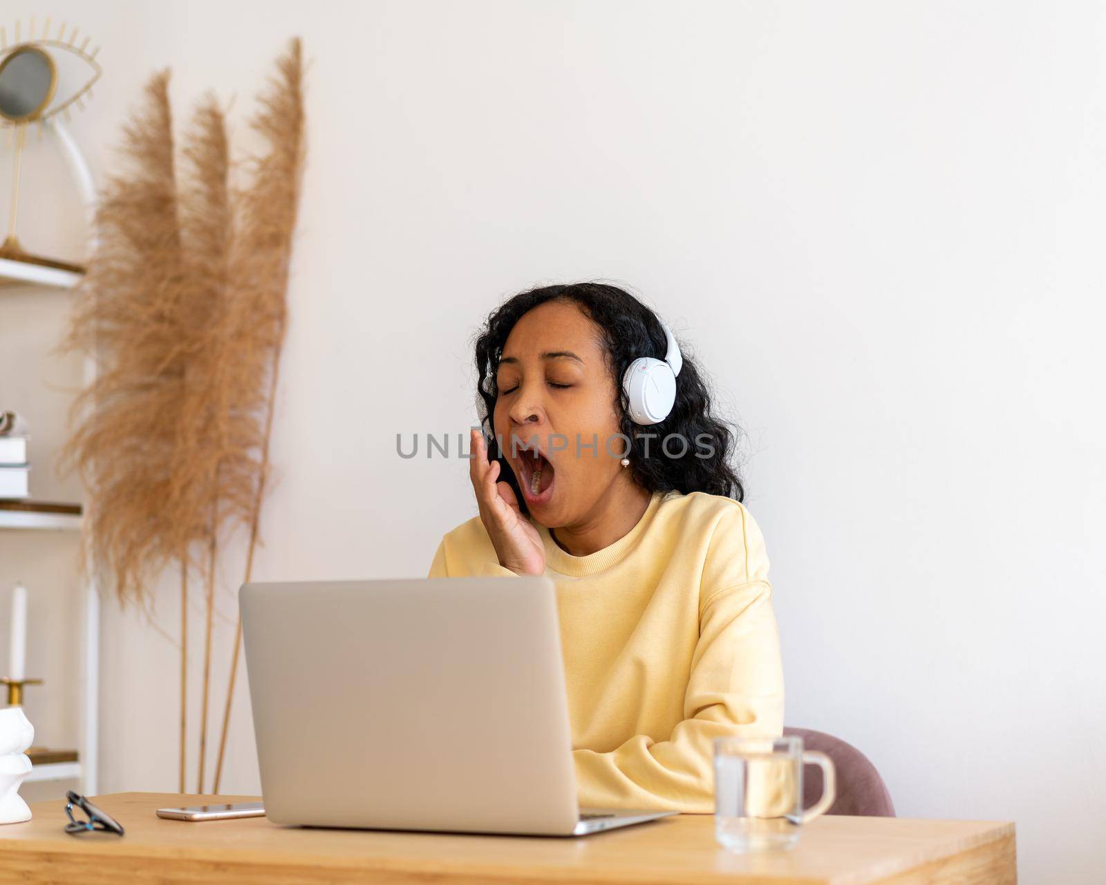 African-american female student yawning while listening to online lecture in headphones on laptop by NataBene
