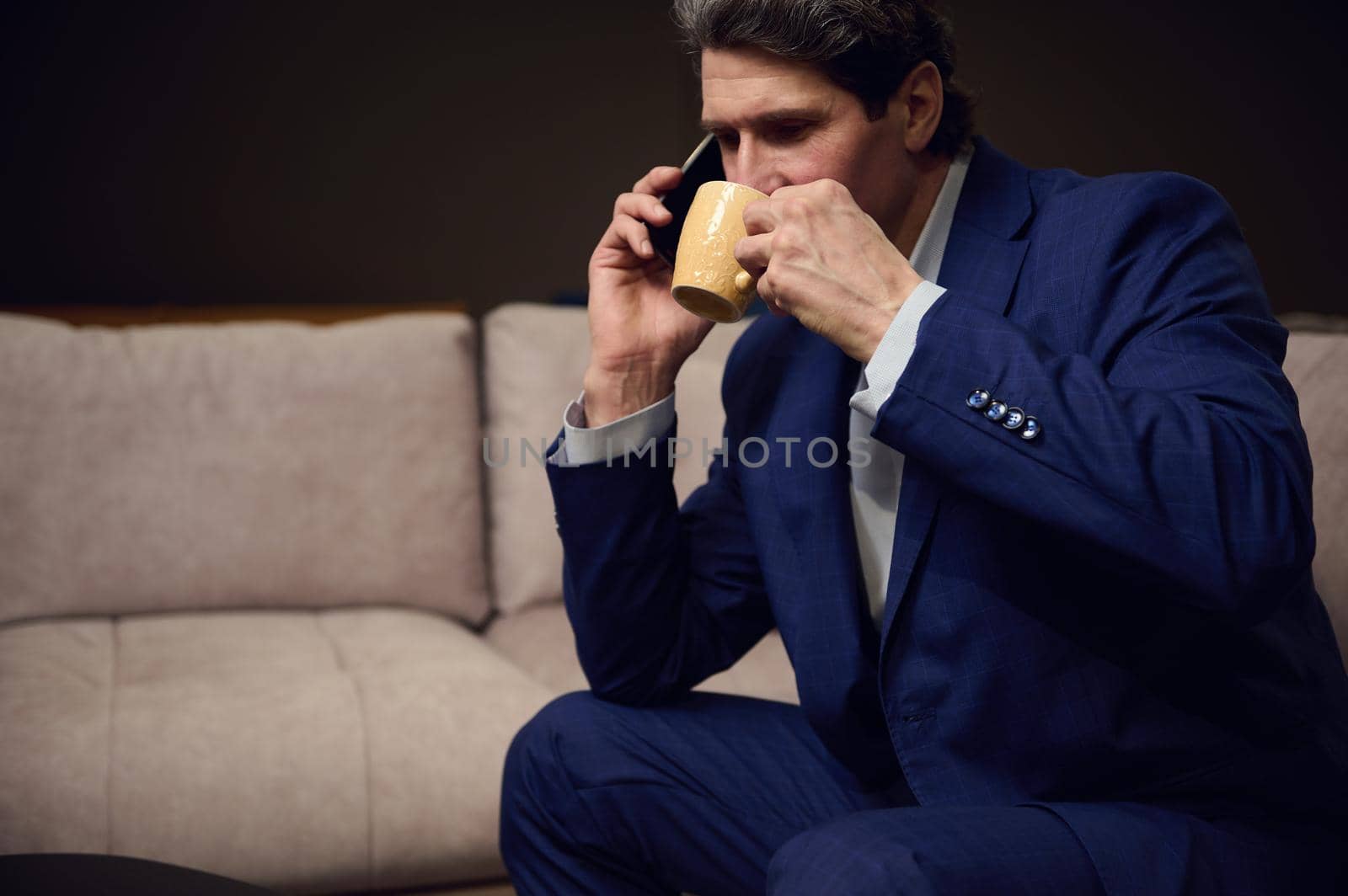 Confident mature businessman, successful entrepreneur, handsome Caucasian man drinking coffee and negotiating on a mobile phone while sitting on a comfortable sofa in his office