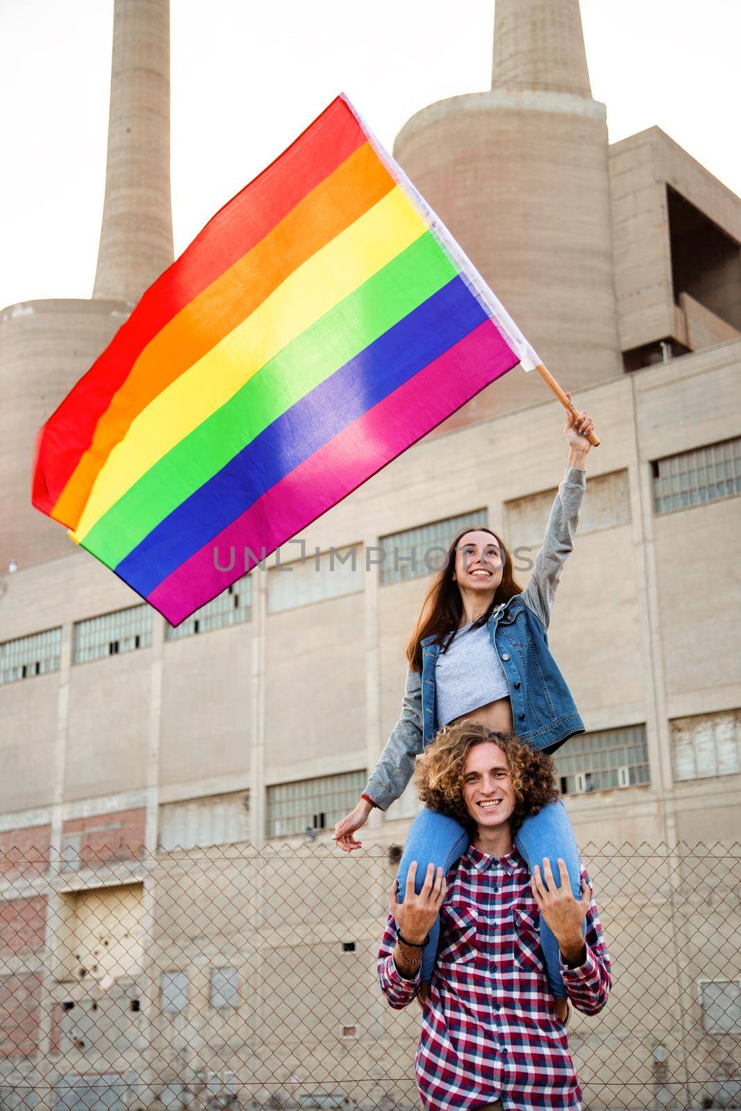 Young happy woman sitting on male friend shoulders waving rainbow flag in LGBT rights demonstration. by Hoverstock