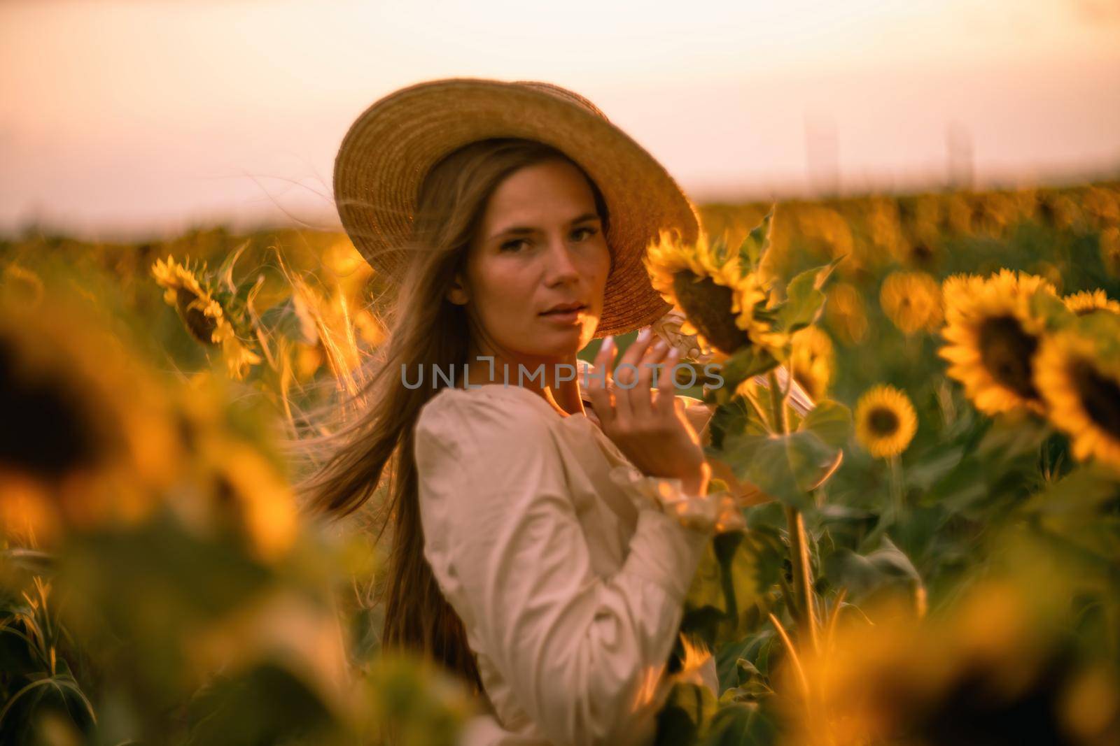 Beautiful middle aged woman looks good in a hat enjoying nature in a field of sunflowers at sunset. Summer. Attractive brunette with long healthy hair. by Matiunina