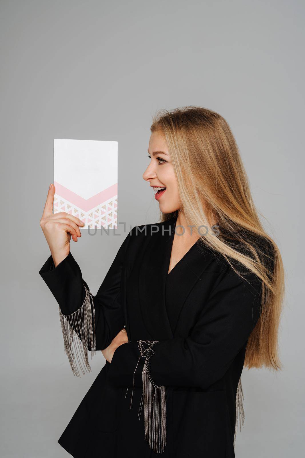 A beautiful middle-aged woman makeup artist holds a diary in her hands and looks at it smiling. Blond hair and a black jacket on a light background. by Matiunina
