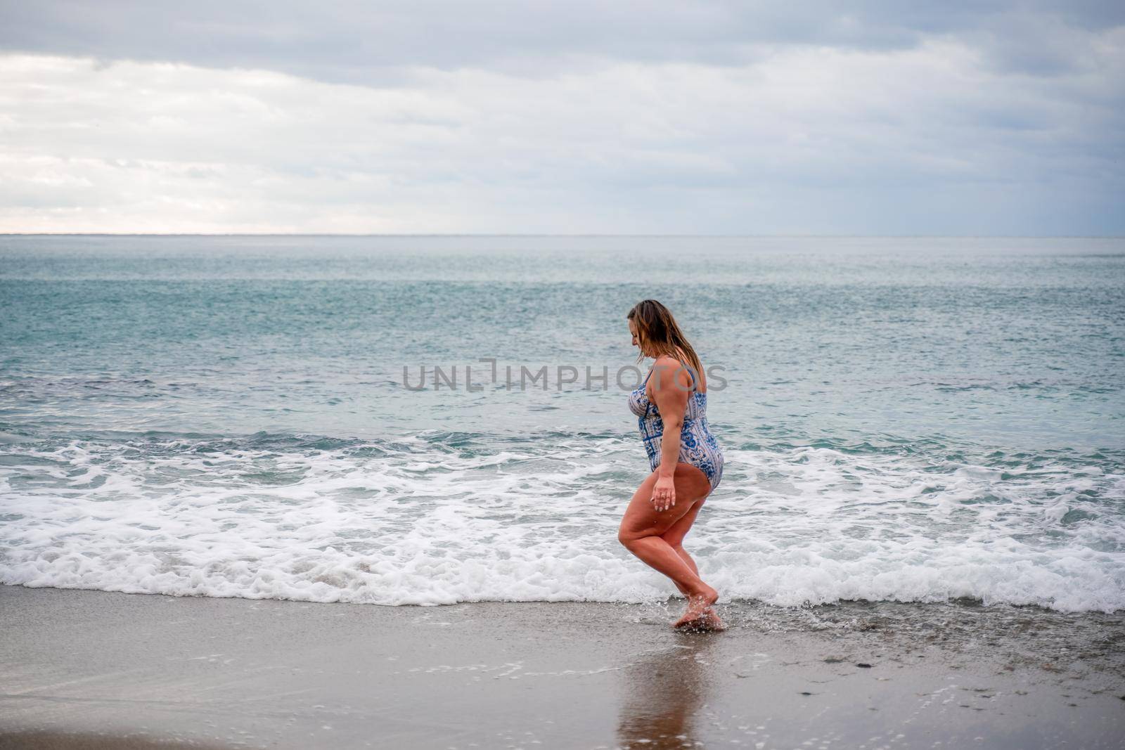 A plump woman in a bathing suit enters the water during the surf. Alone on the beach, Gray sky in the clouds, swimming in winter. by Matiunina