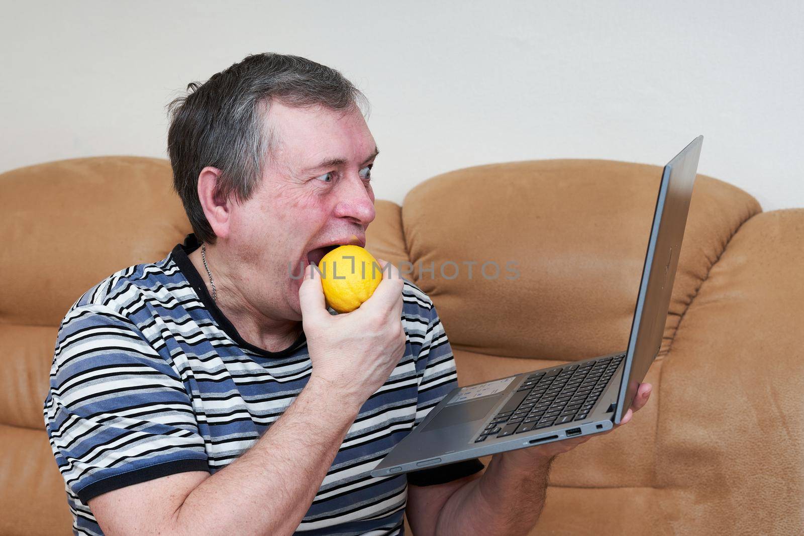 Freak eats lemon while holding a laptop while sitting on the couch. Emotions from the news