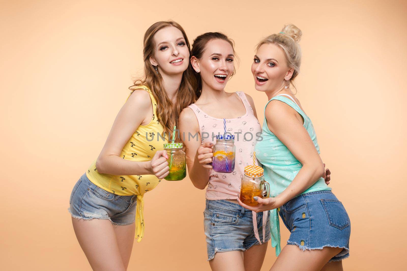 Three beautiful slender women in summer clothes with cocktails are laughing, having a good time together. Studio, isolated background. The concept of recreation