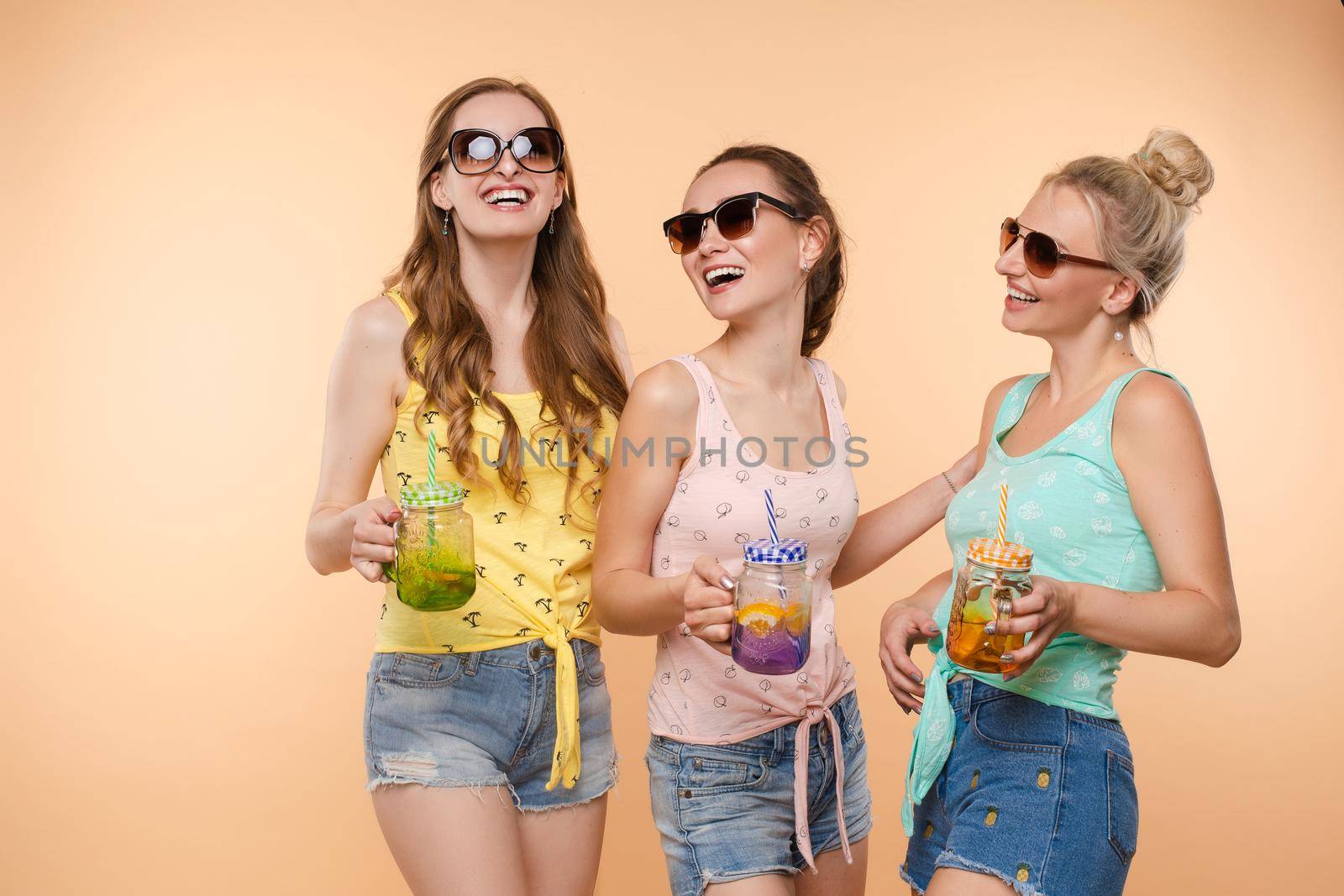 Three beautiful slender women in summer clothes with cocktails are laughing, having a good time together. Studio, isolated background. The concept of recreation