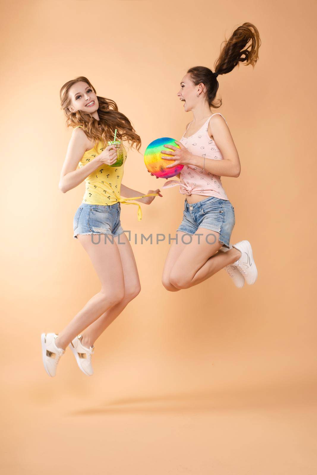 Three beautiful slender women in summer clothes with cocktails are laughing, having a good time together. Studio, isolated background. The concept of recreation